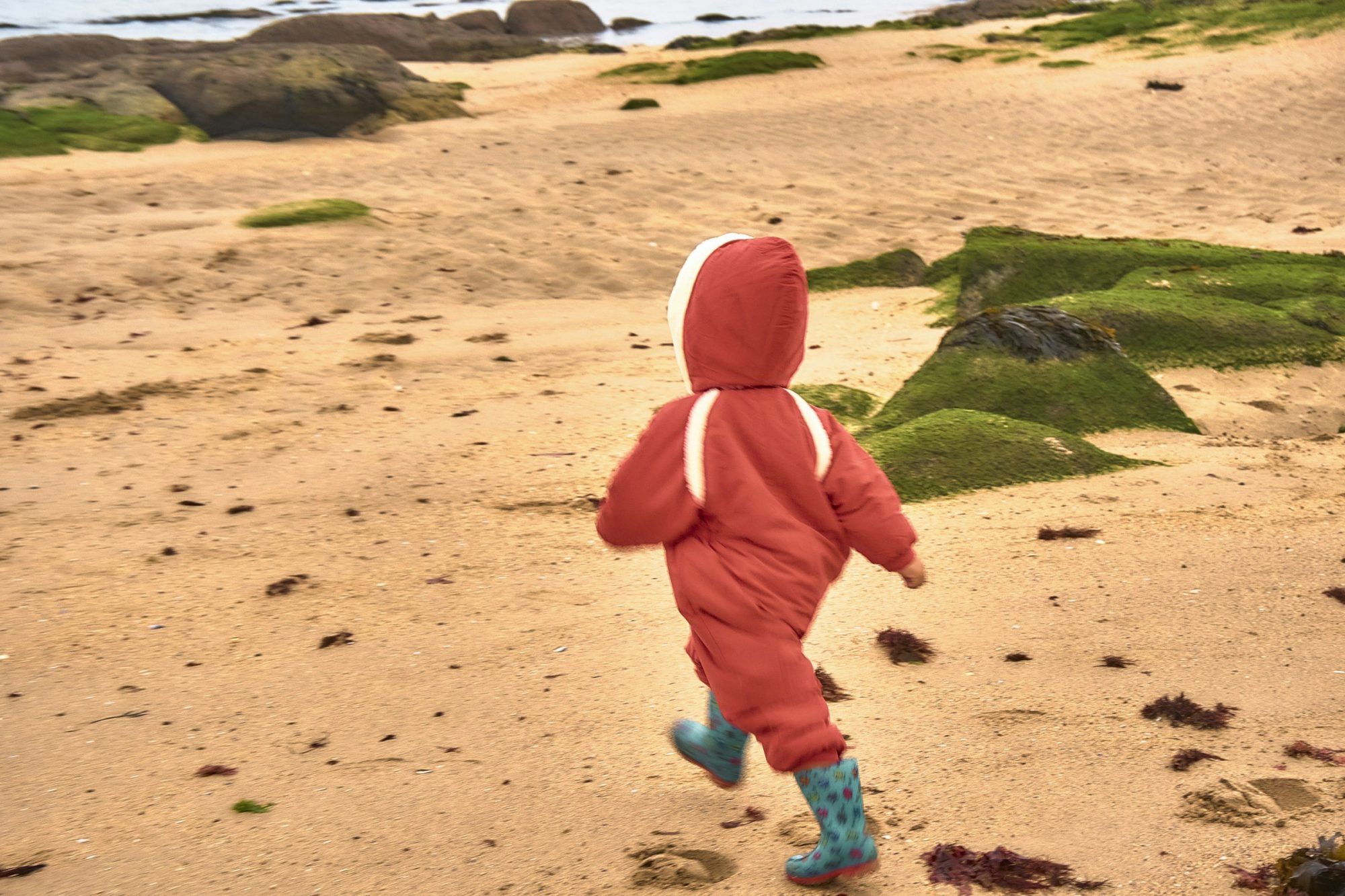 Un bambino con la tuta rossa corre di spalle su una spiaggia di sabbia.