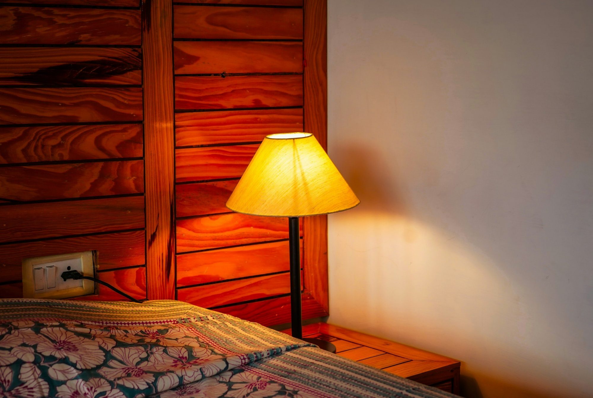 Una lampada da comodino accesa diffonde una luce calda accanto a un letto con la coperta a fiori