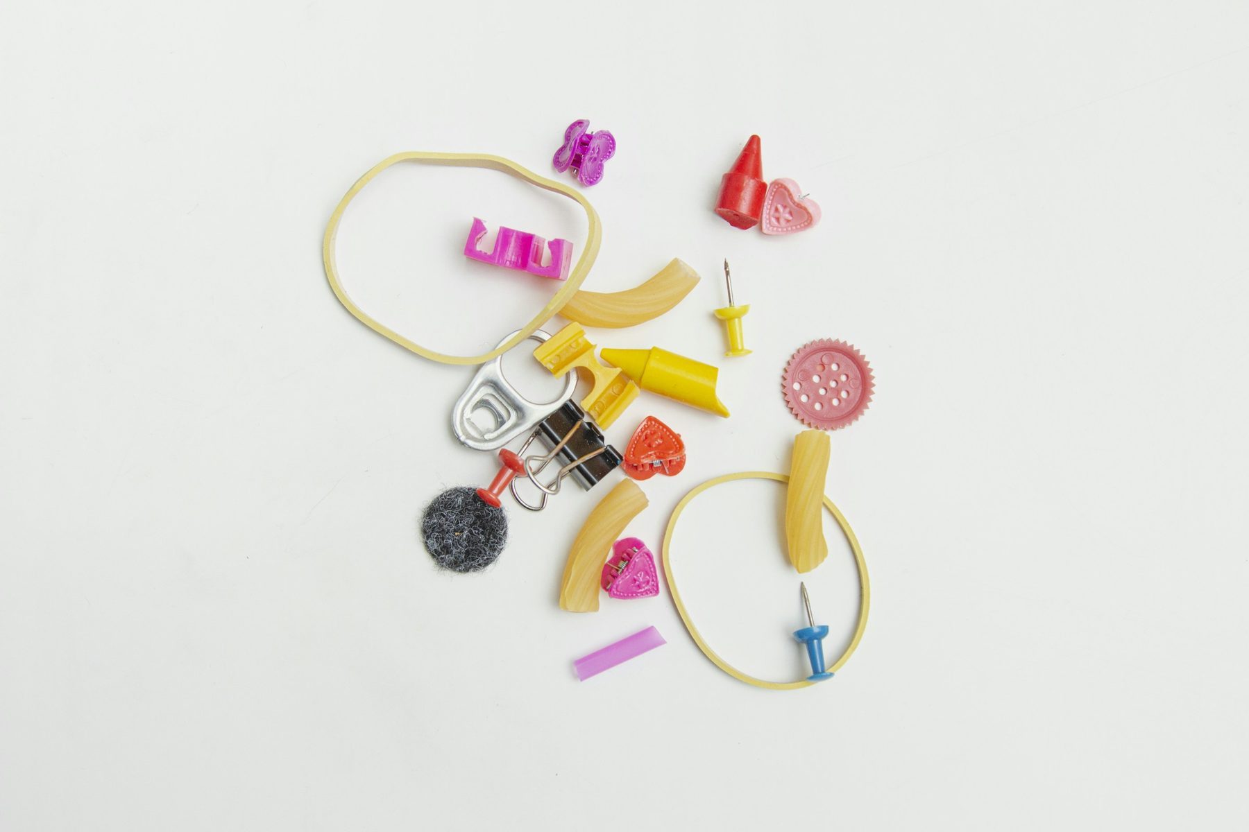 Piccoli oggetti colorati sparsi su un piano bianco, tra fermagli a forma di cuore, elastici, puntine e mollette