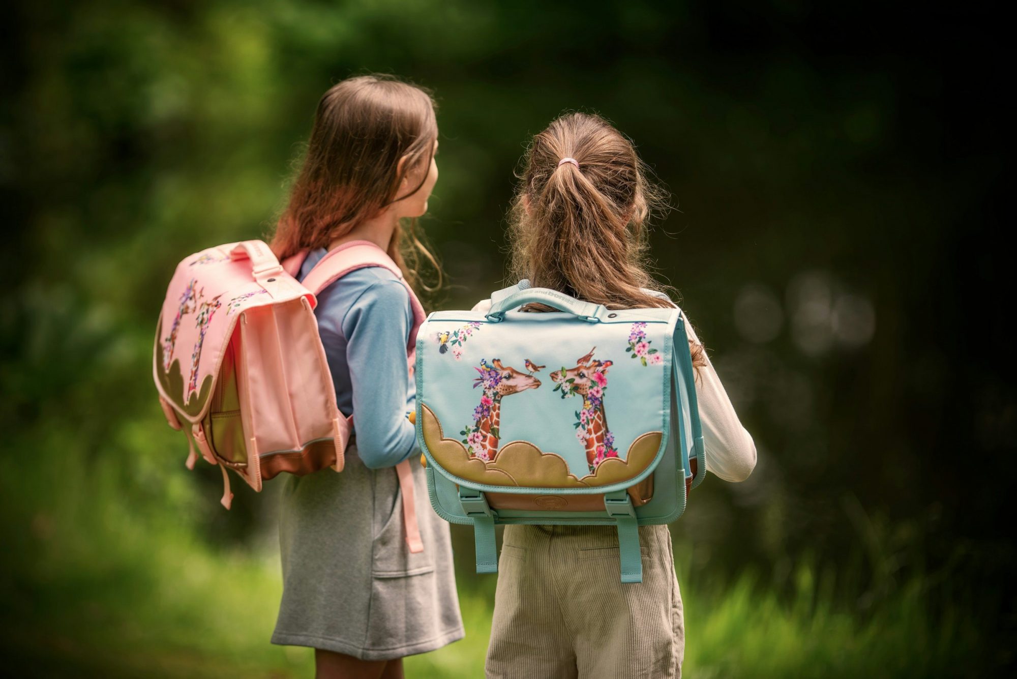 Due bambine viste di spalle con gli zaini di scuola, immerse nel verde.